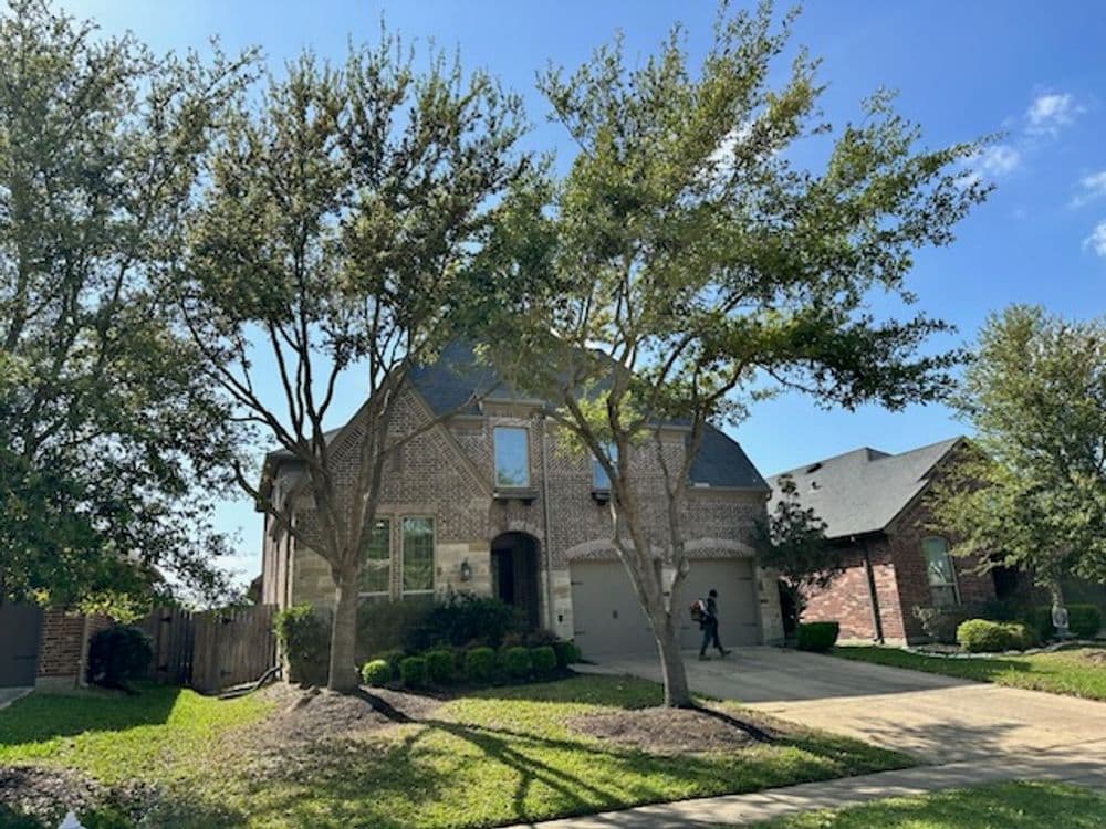 Brick house with trees, blue sky, and a person walking by on a sunny day.