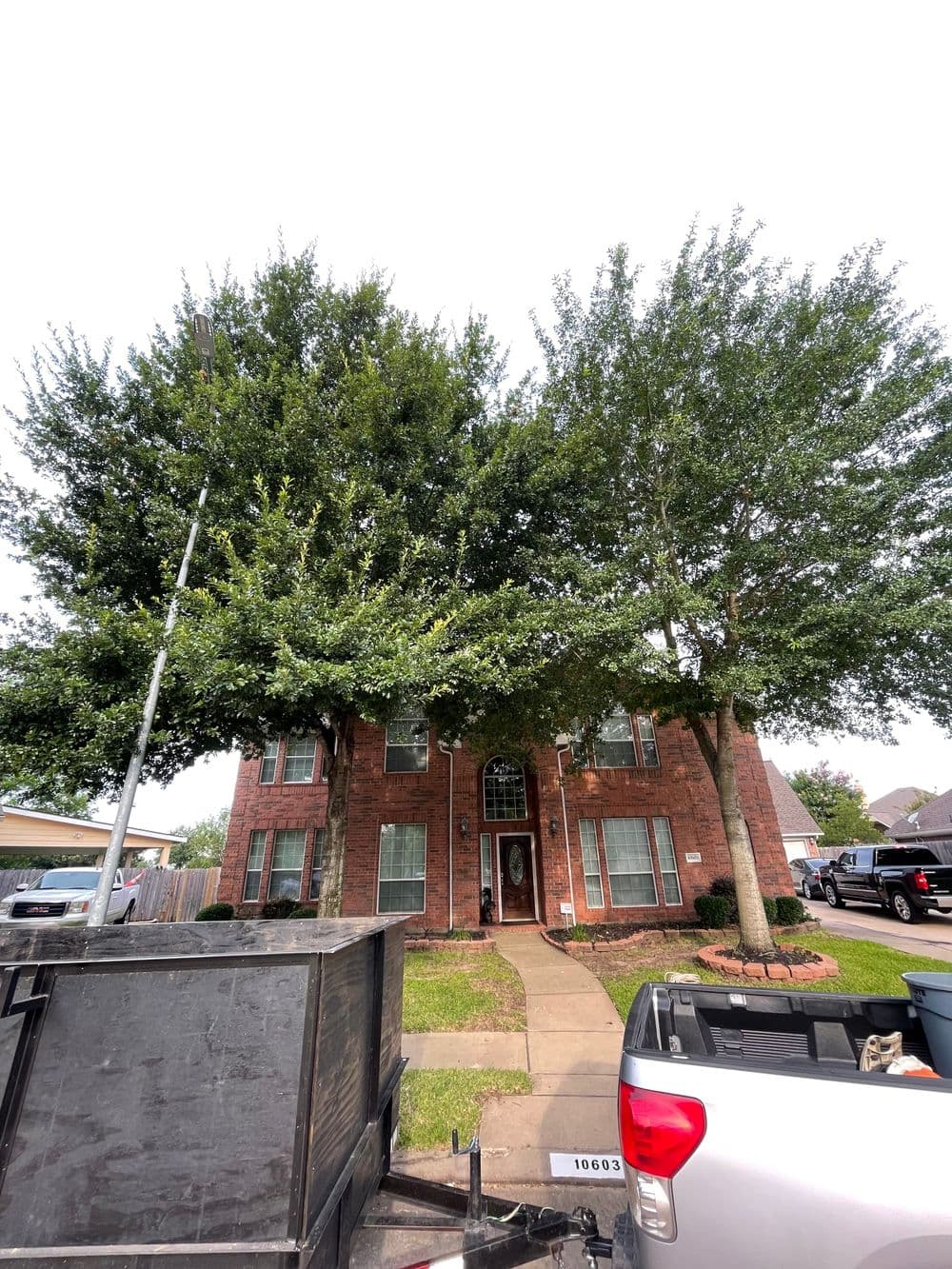 Two-story brick house with large trees in front and landscaping on a sunny day.