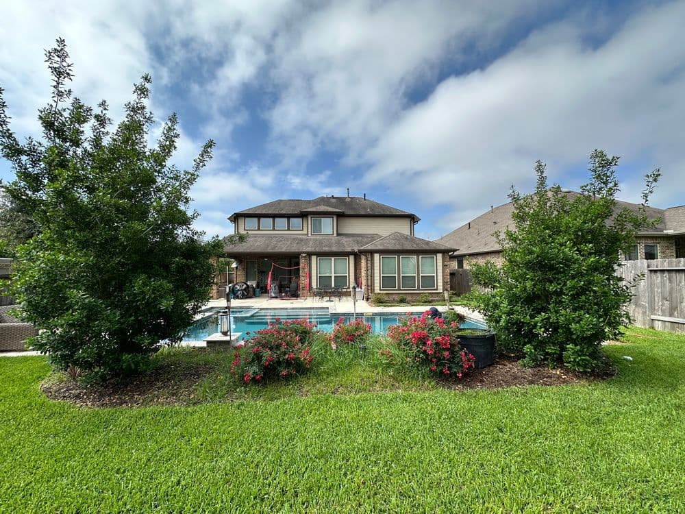 Backyard with lush greenery, blooming flowers, and a pool, under a partly cloudy sky.