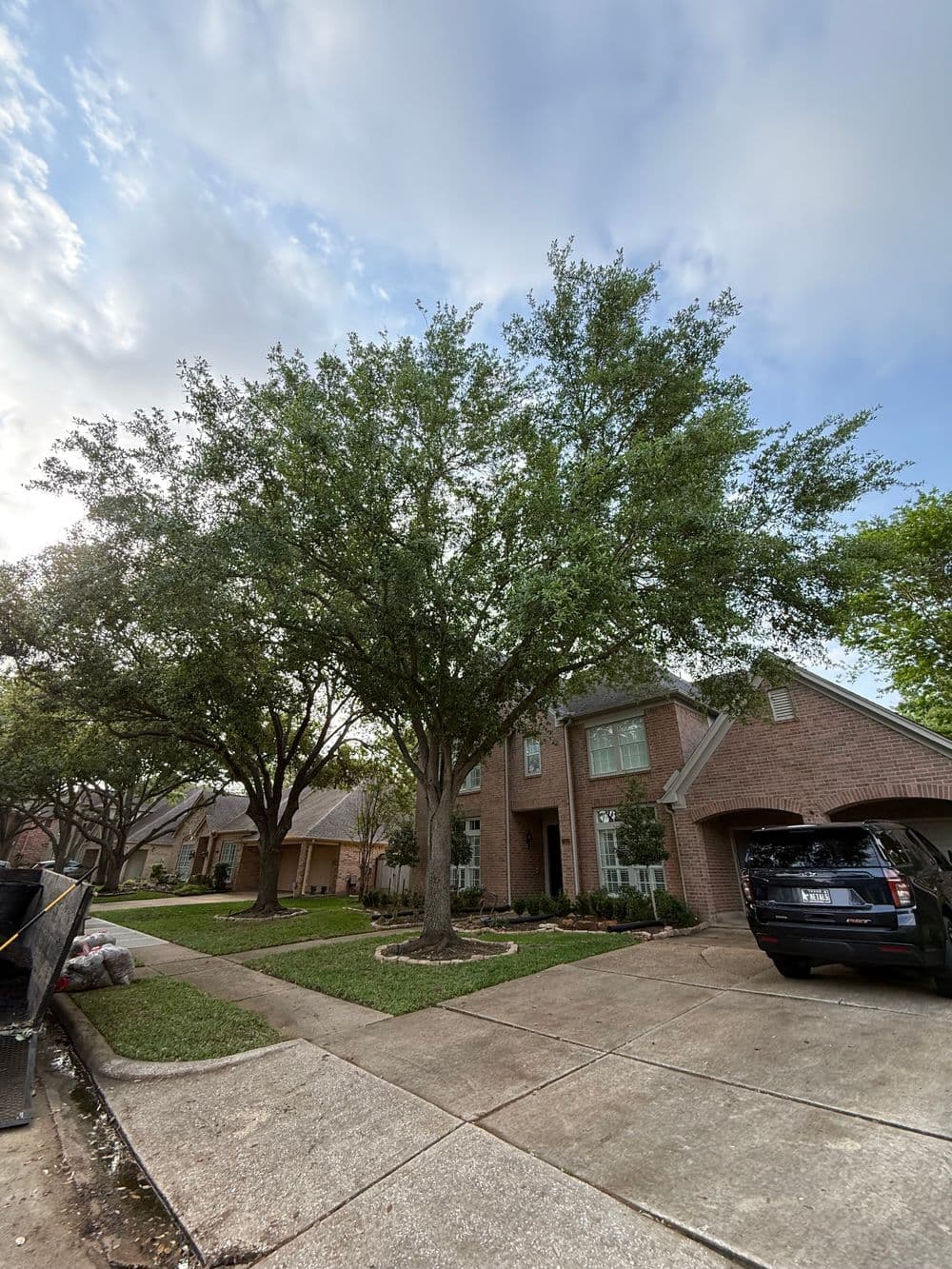 Brick house with large trees and well-maintained lawn, car parked in driveway. Clear sky.