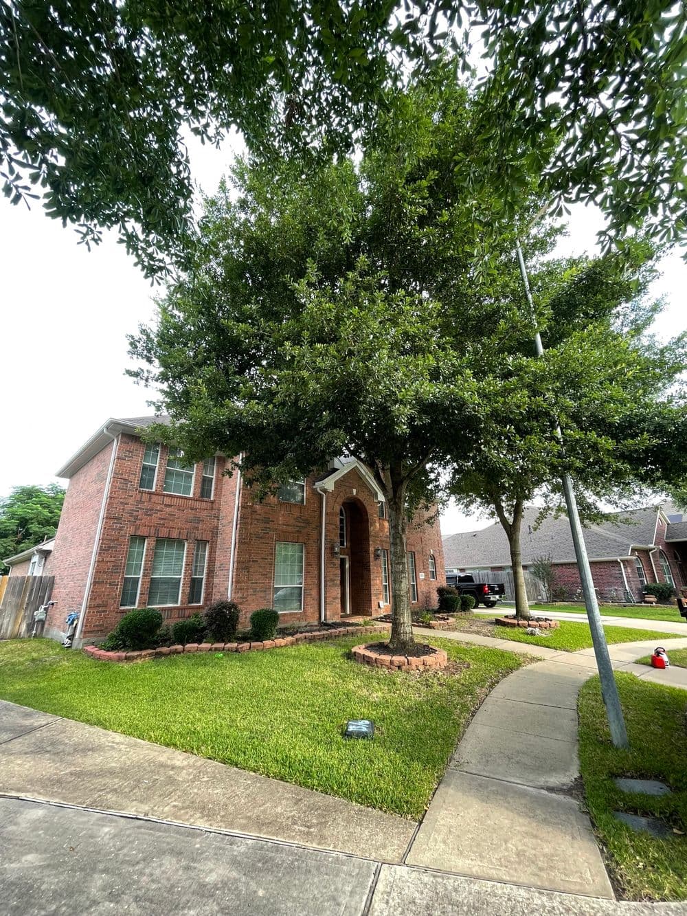 Brick house with tall trees and landscaped yard, featuring a curved sidewalk in a suburban setting.
