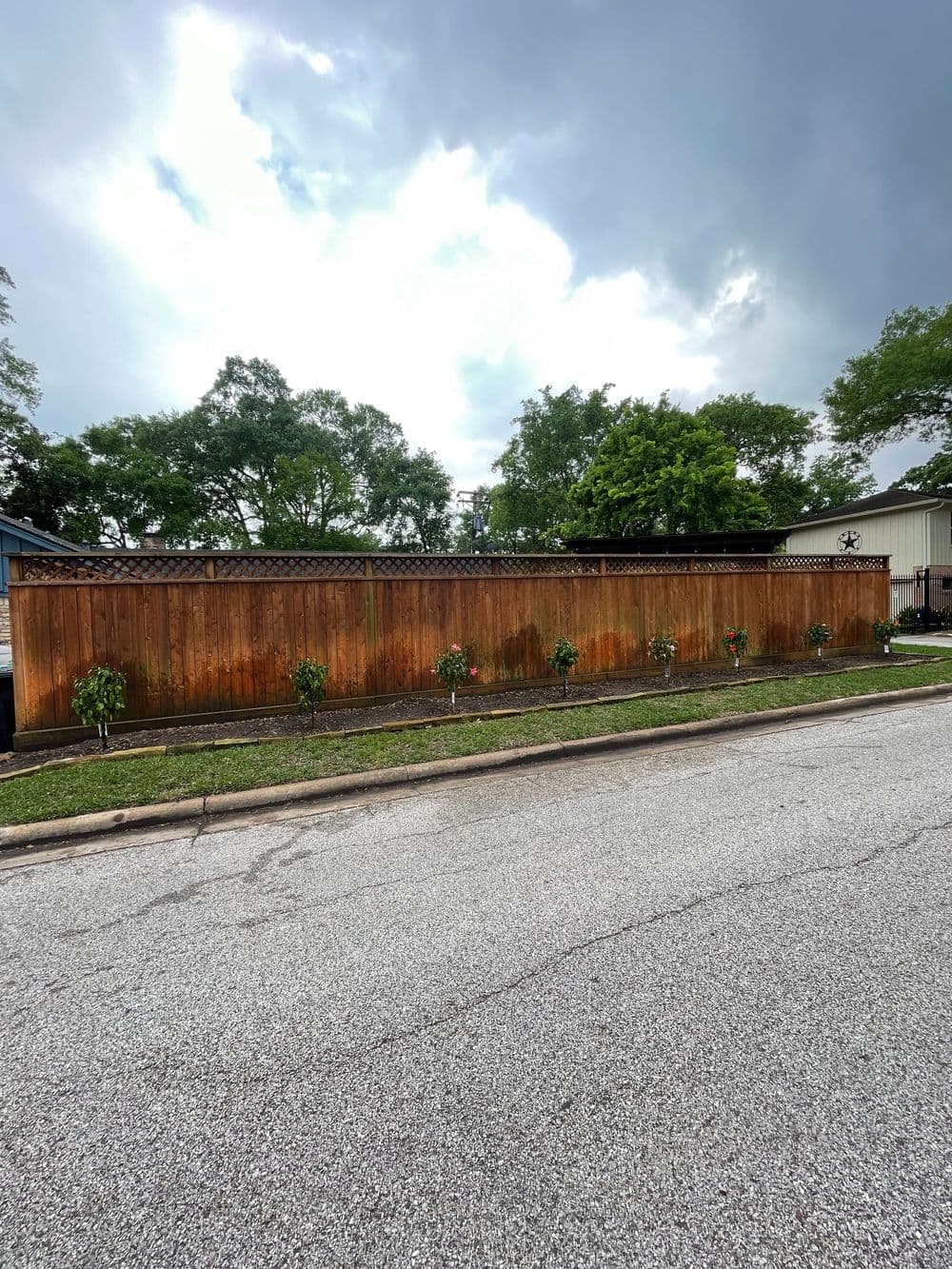 Wooden fence with landscaping and small shrubs along a residential street under cloudy sky.