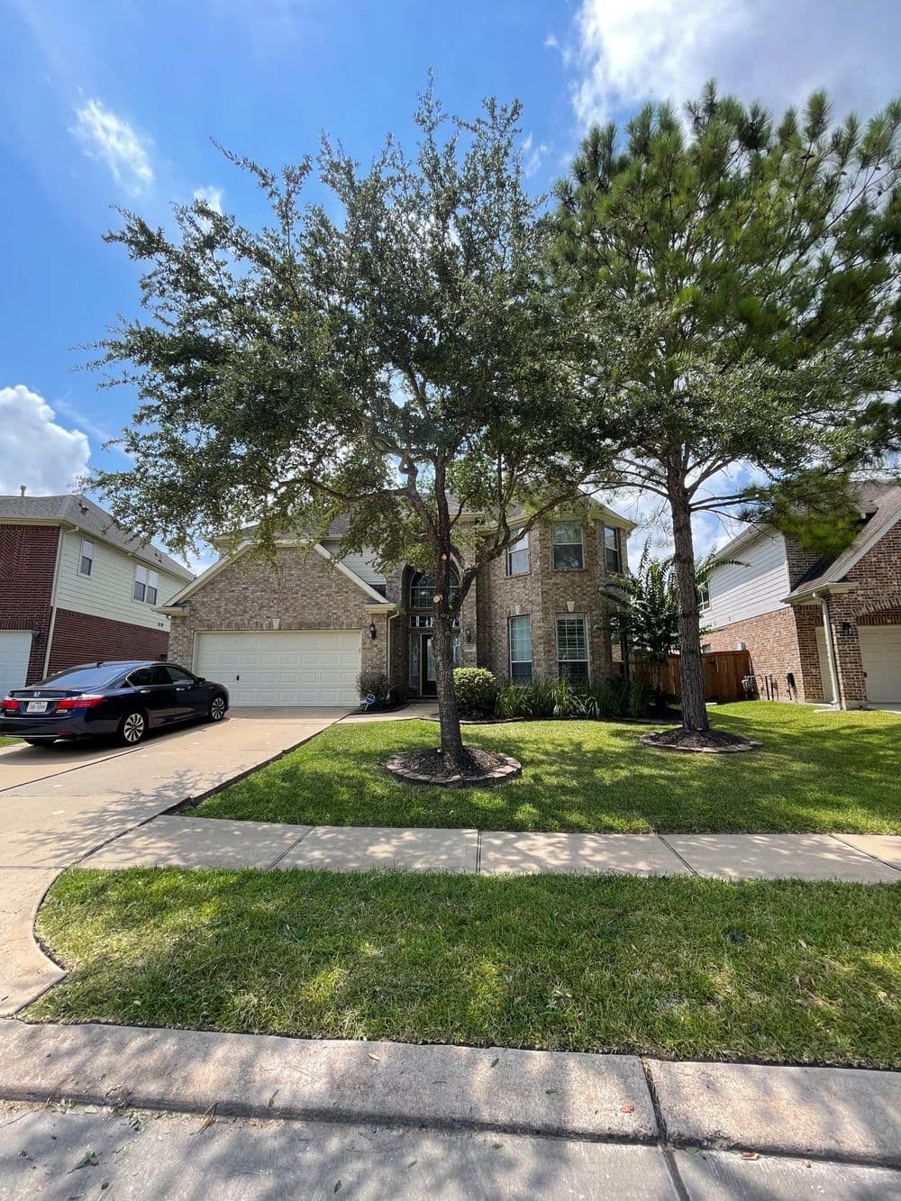 Two large trees shade a suburban home with a well-maintained yard and driveway.