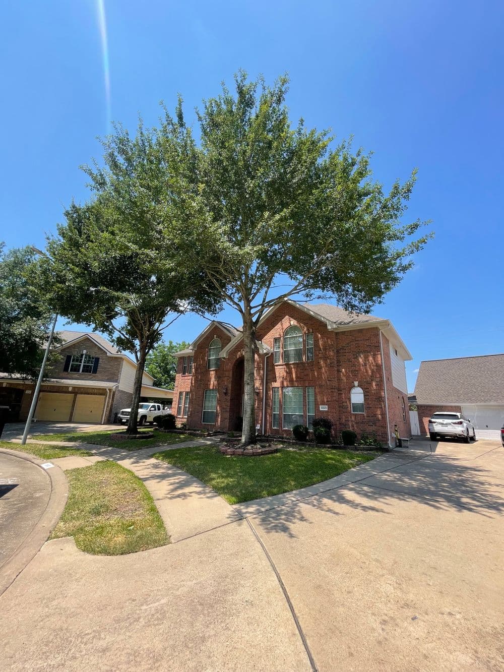 Two-story brick house with large trees in front, sunny blue sky, and driveway visible.